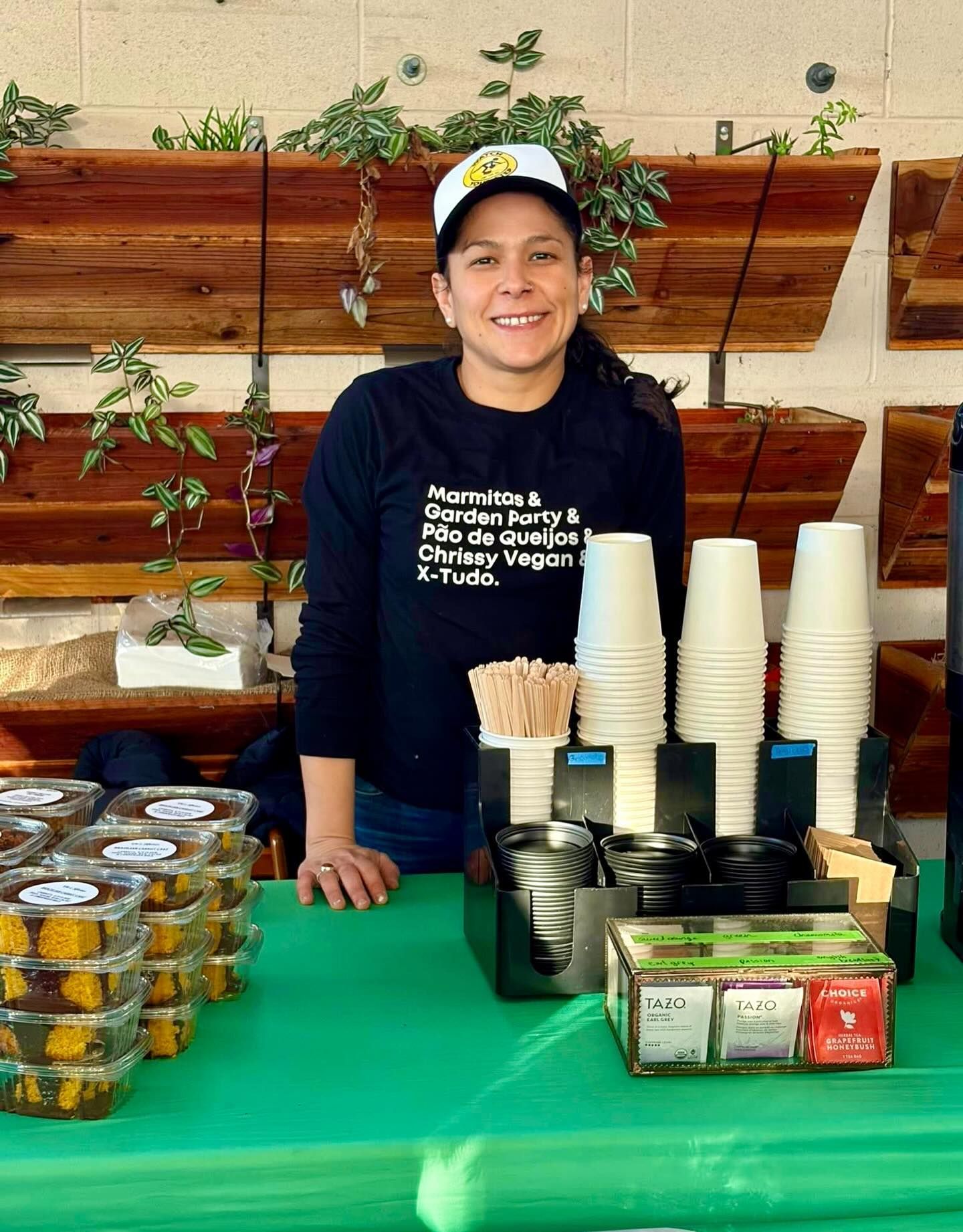 Smiling vendor behind a green-covered table with stacked cups, napkins, and food containers at an indoor booth