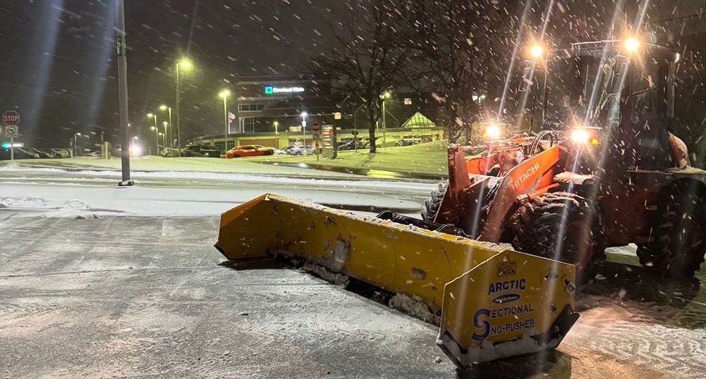 Orange snowplow clearing a snowy parking lot at night under streetlights