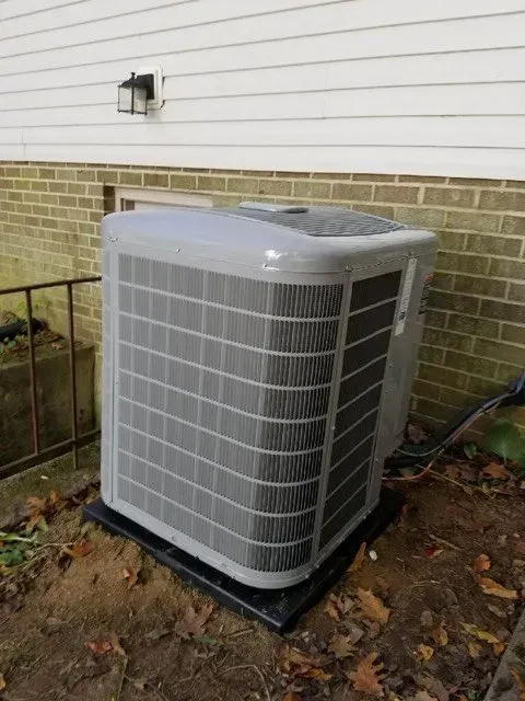 Gray air conditioning unit outside a building with white siding and brick.
