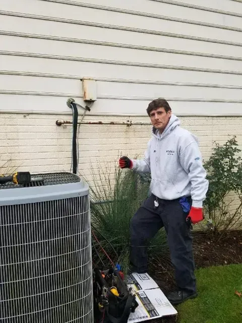 Man in work clothes near an air conditioner unit by a house. He points at the unit.