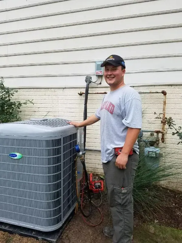 Man in work clothes smiles next to an air conditioning unit outside a house.