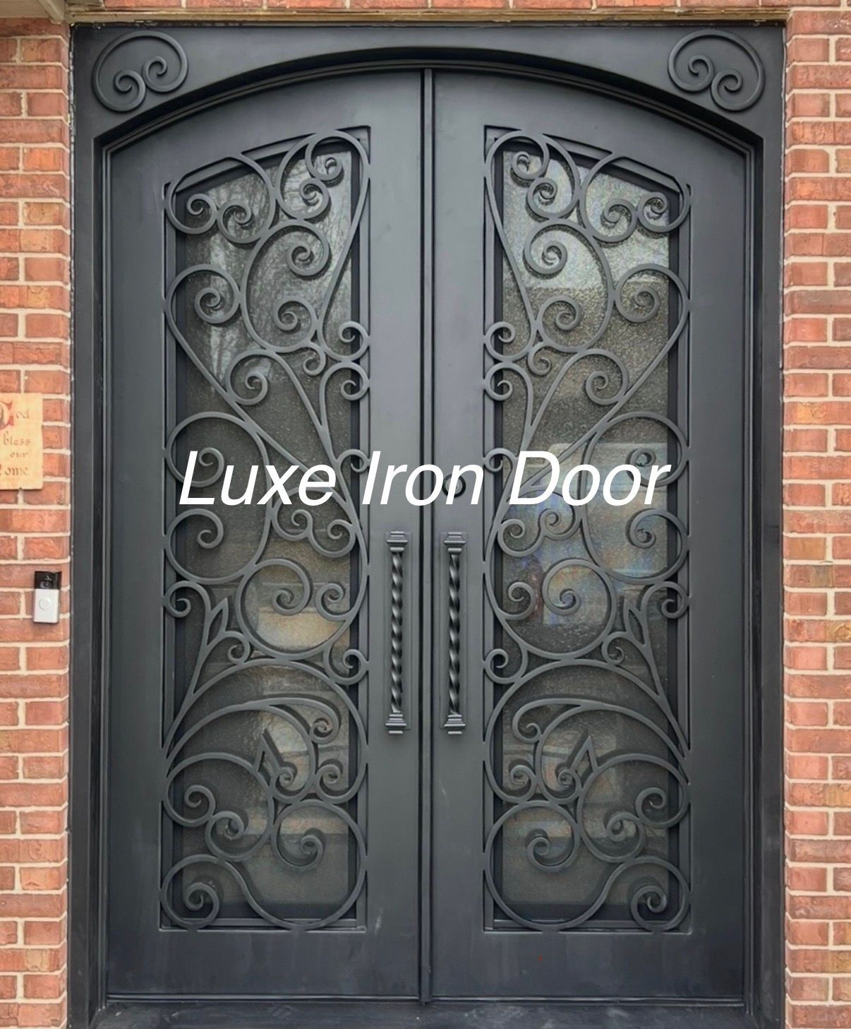 A black arched double iron door with intricate scrollwork, set against a red brick wall.
