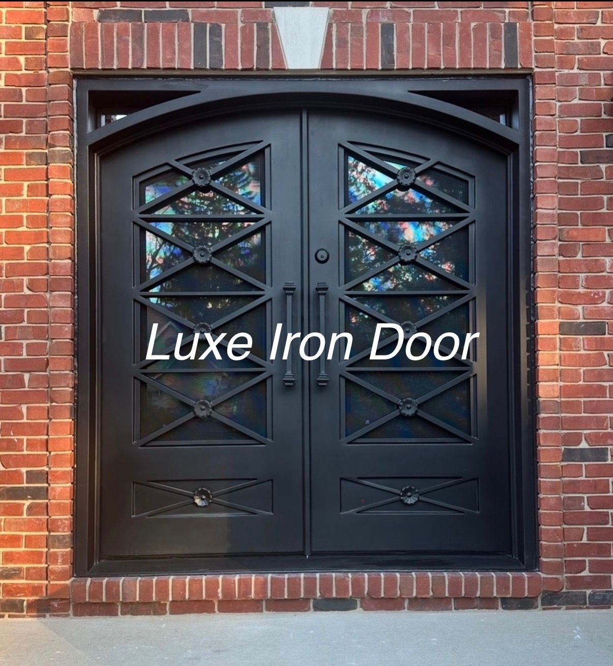 A double black iron door with arched top glass panels and geometric metal accents, set within a red brick entryway.