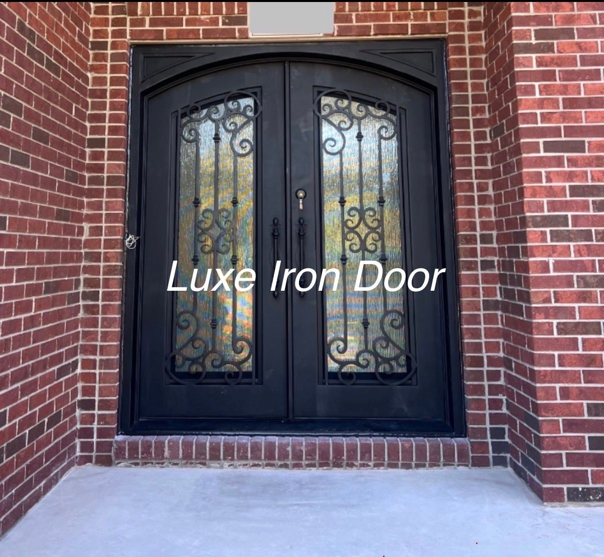 A black double iron arched entry door with decorative scrollwork glass inserts, set into a red brick house facade.