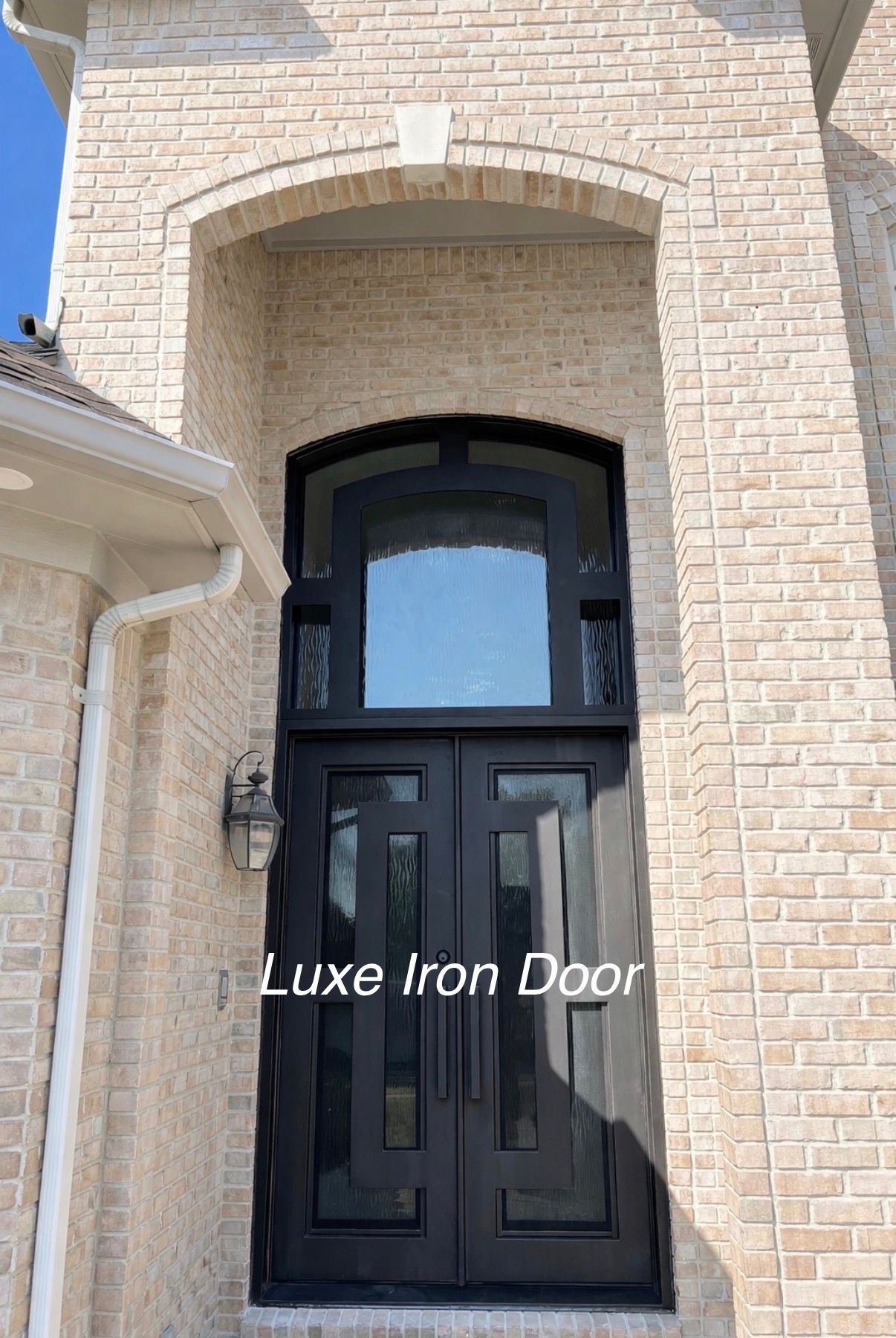 A modern black iron double door with a rectangular glass design and arched transom window on a light brick house exterior.
