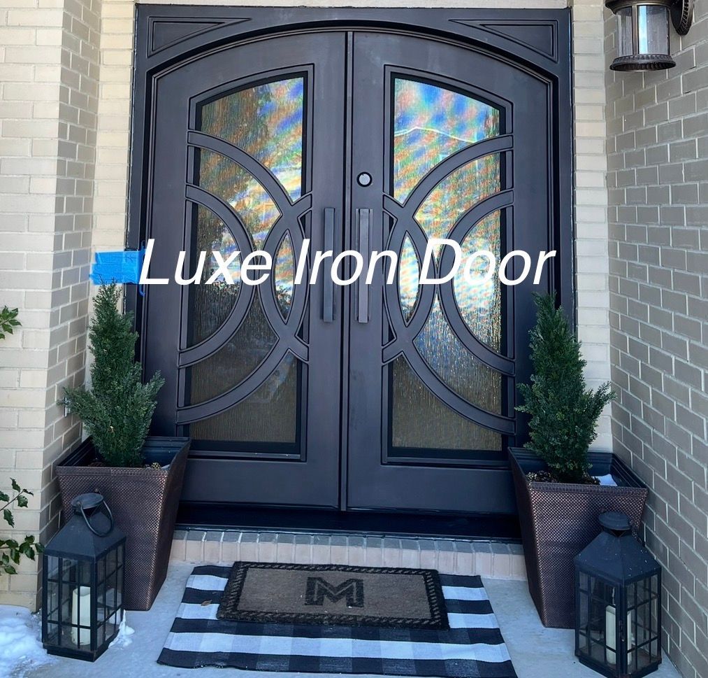 A black double iron front door with arched glass patterns, flanked by two potted evergreens and lanterns on a porch.
