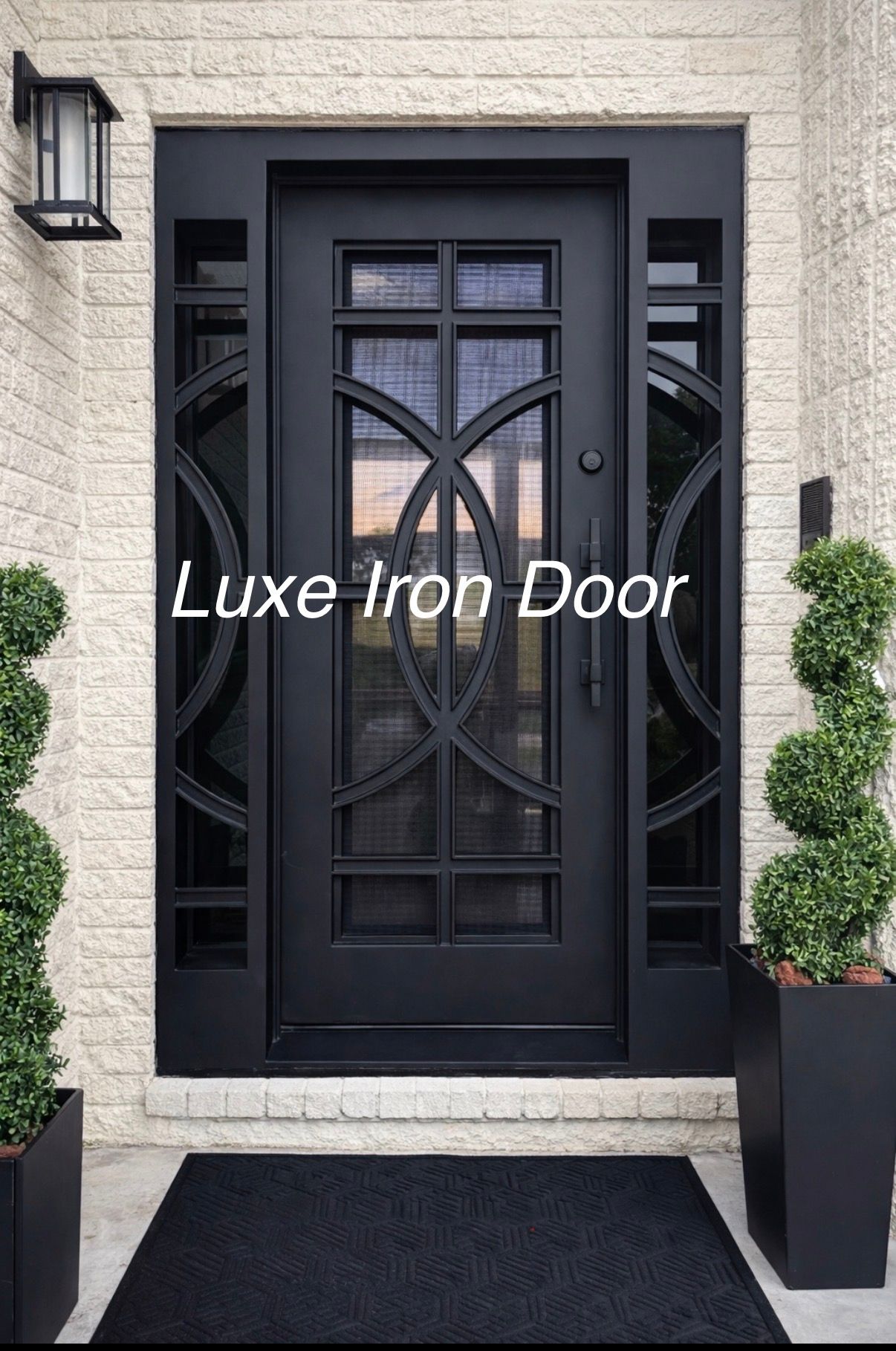 Black iron front door with glass geometric inserts and sidelights, flanked by two spiral topiaries on a brick porch.