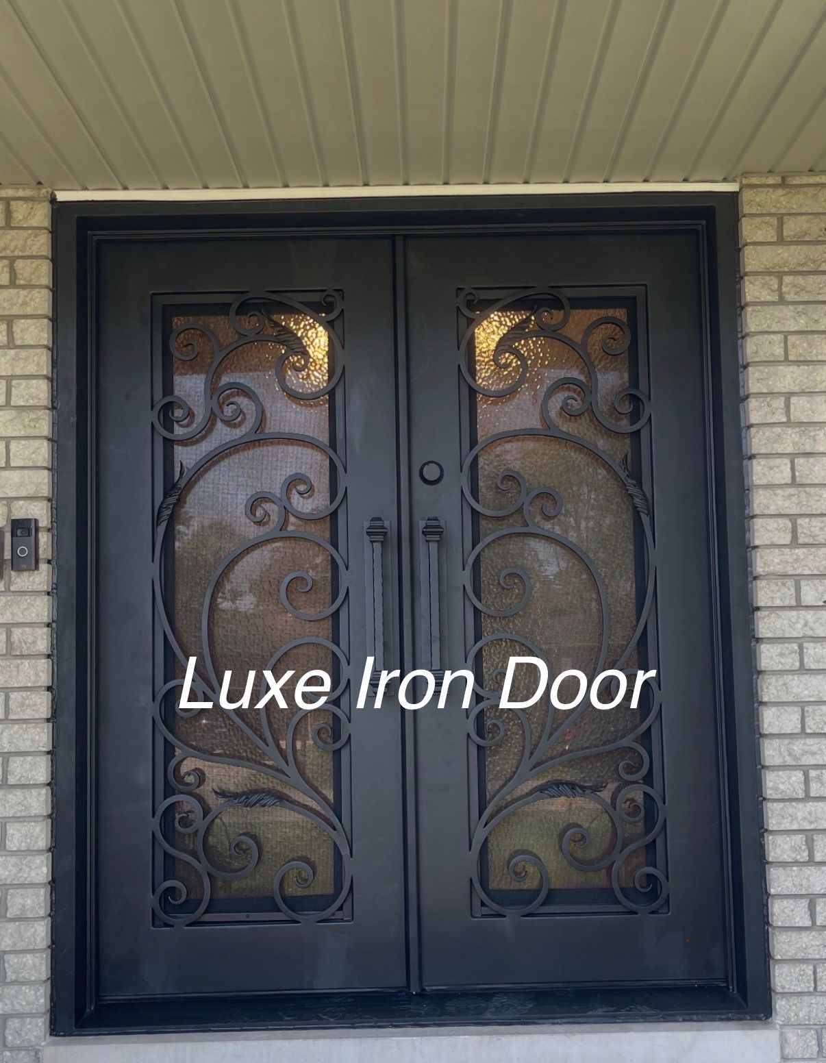 Black double wrought iron doors with ornate scrollwork over glass panels, set in a light brick wall.