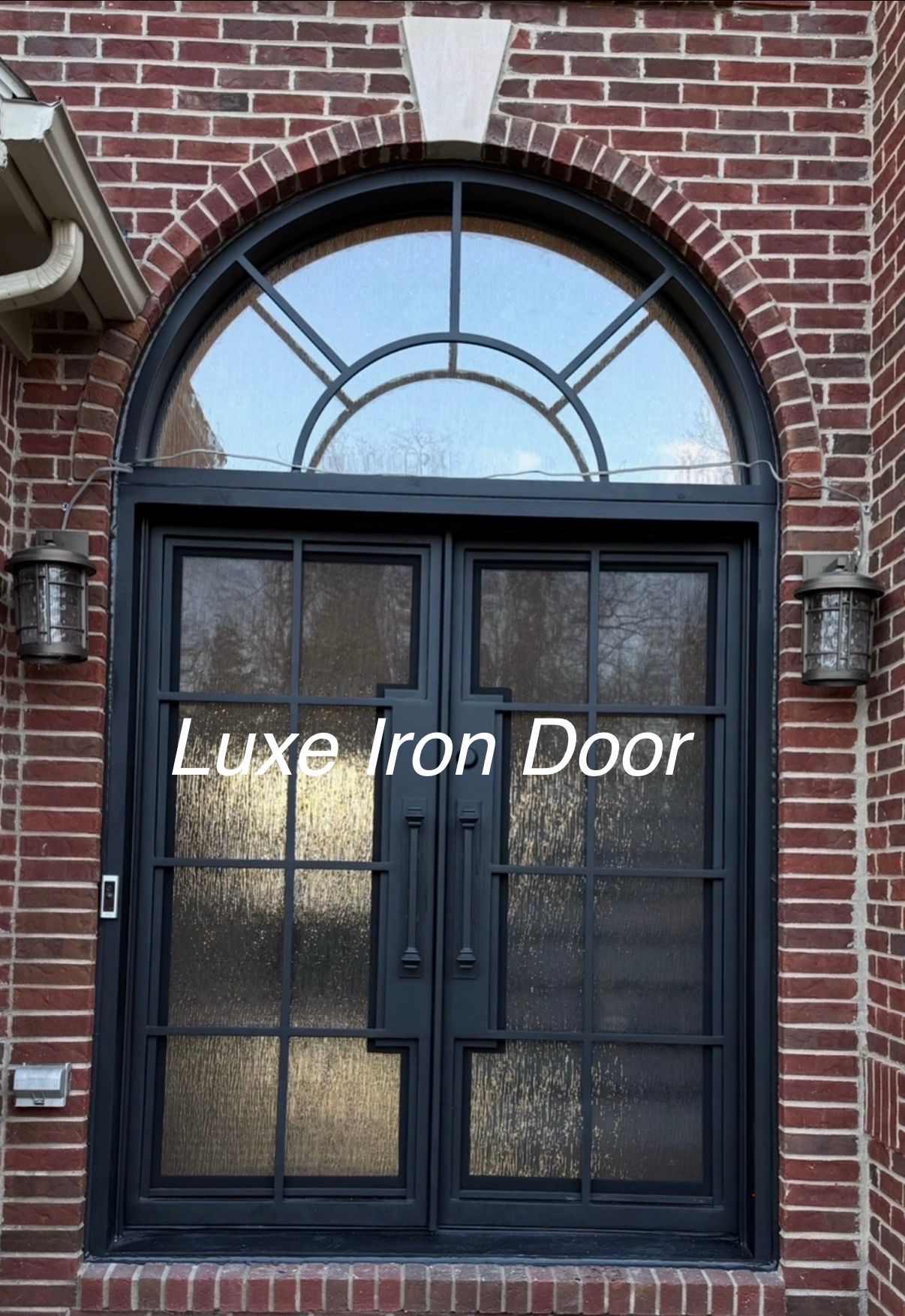Black arched double iron door with textured glass panels and a transom window, set in a brick exterior wall.