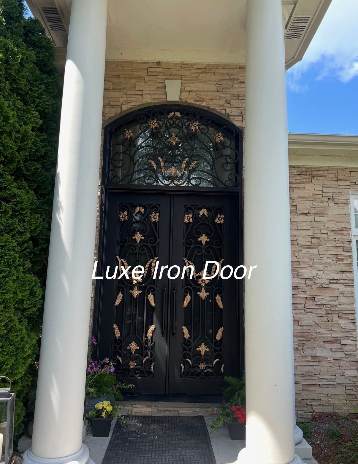 Double iron front door with ornate gold detailing, arched glass transom, and white columns on a stone porch.