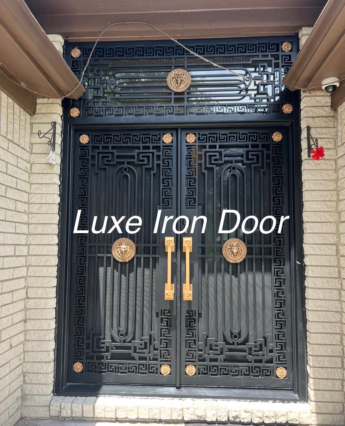 A black iron double door with geometric Greek key patterns, gold-colored lion head accents, and vertical gold handles.