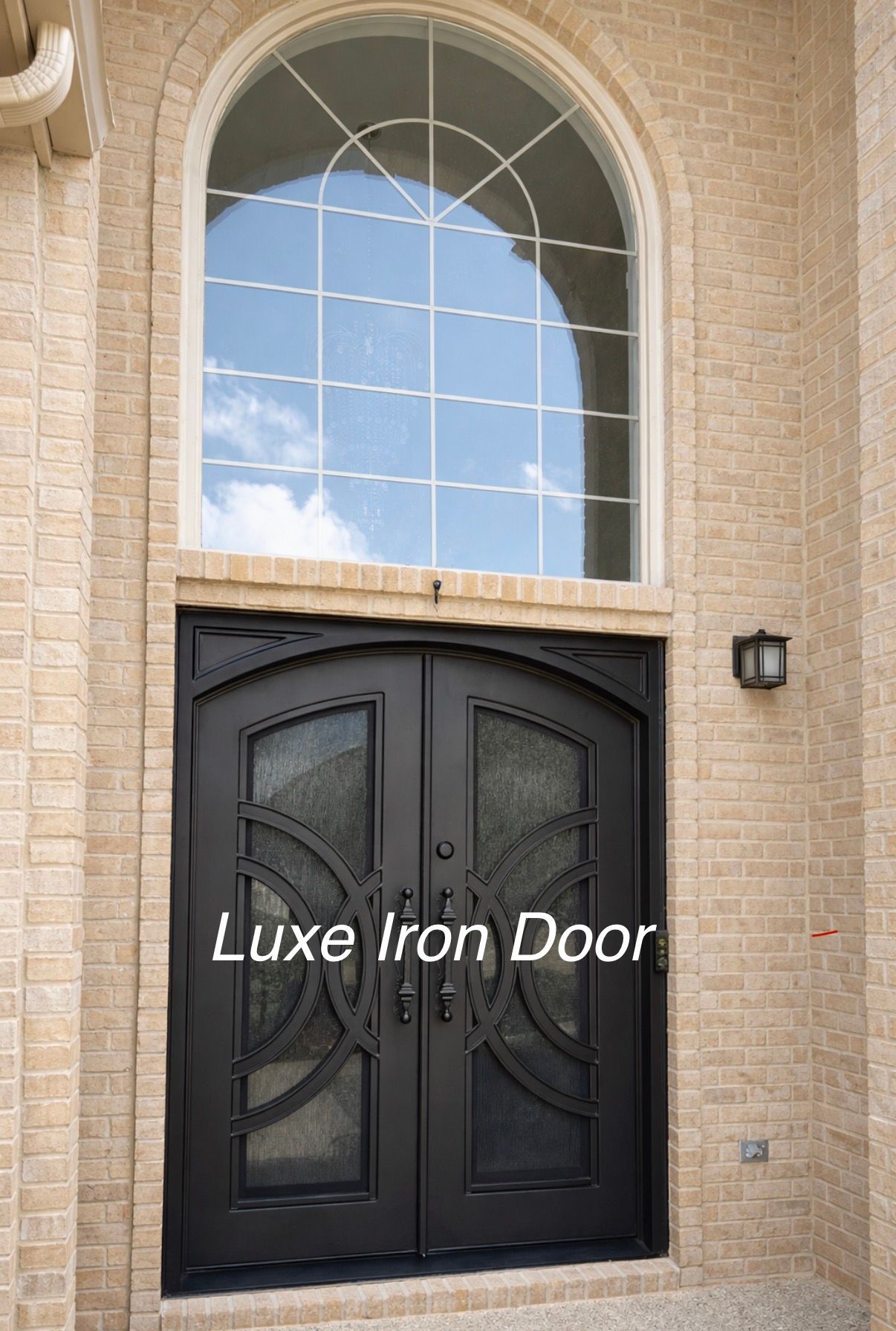 Black double iron front door beneath a large arched window on a brick house facade
