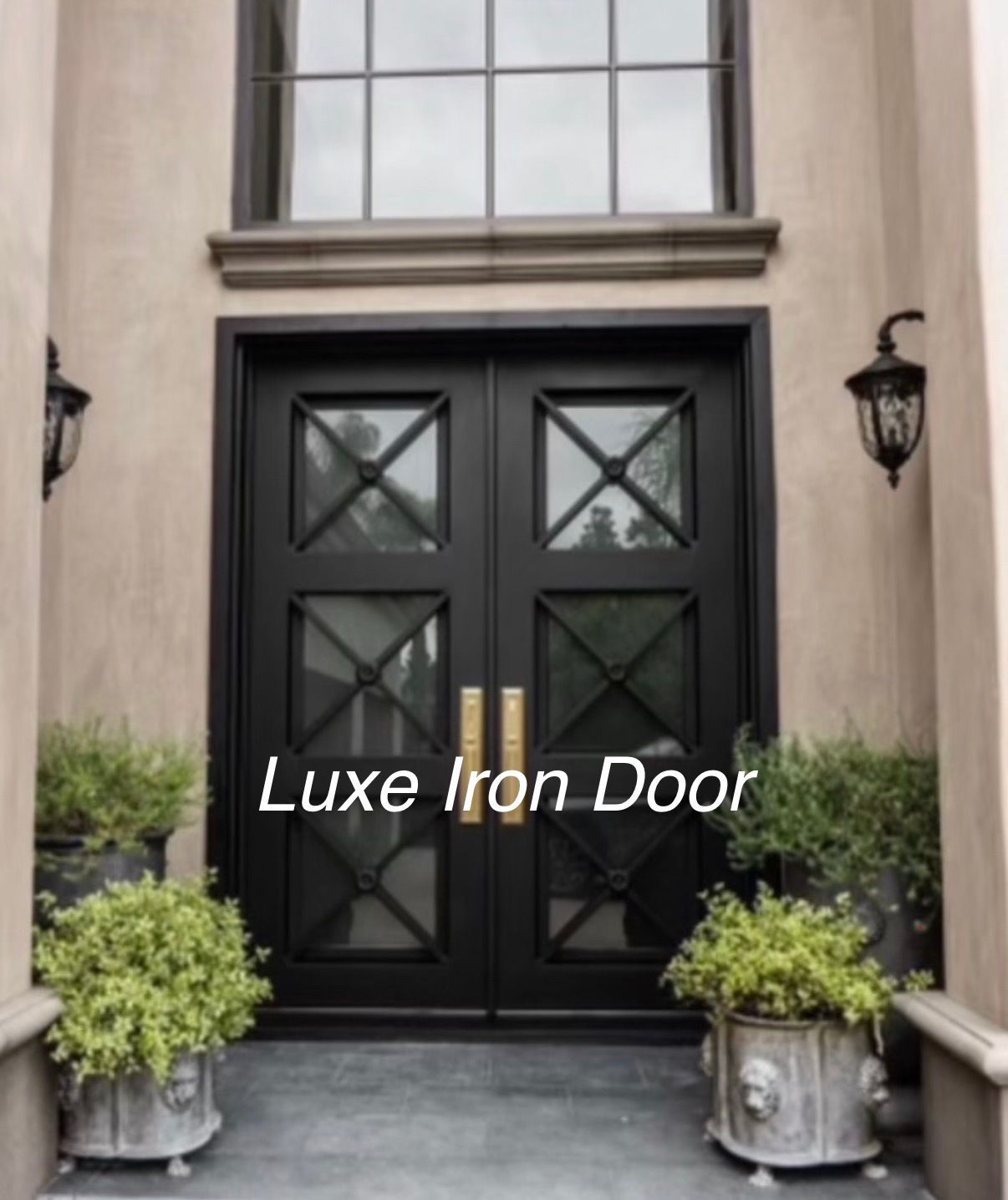 Black iron double front door with decorative glass panels, flanked by wall lanterns and potted shrubs.
