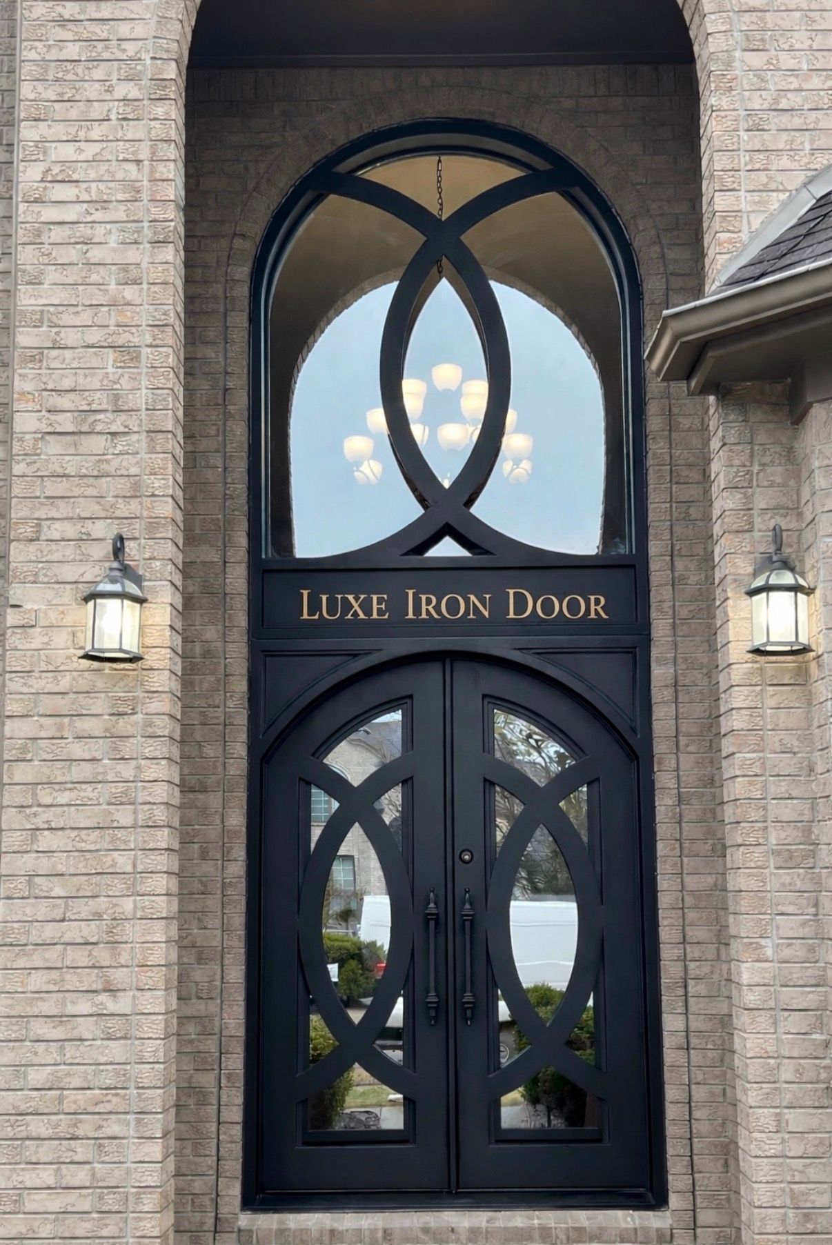 Black luxury iron double door with arched transom in a stone entryway, flanked by wall lanterns and planters