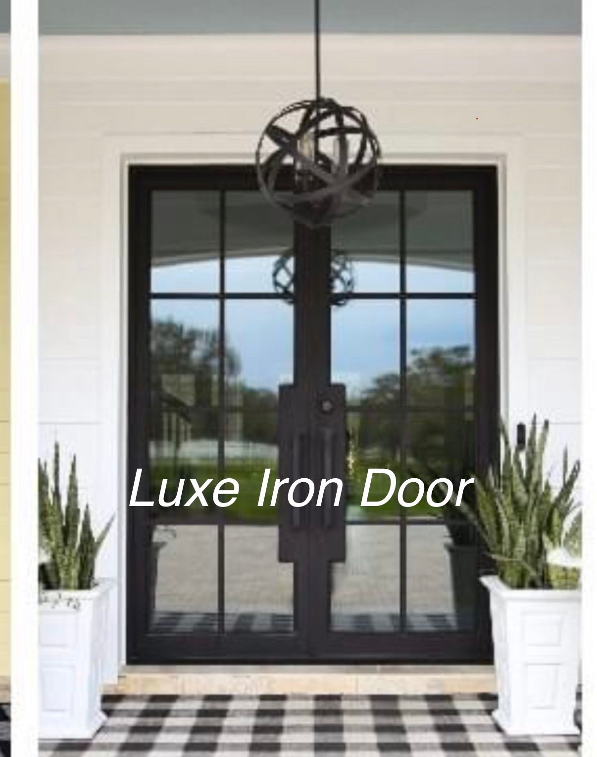 Black iron double front door with glass panes, centered beneath a decorative metal wheel and flanked by white planters.
