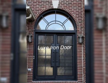 A black double iron door with a glass arched transom and textured glass panels, framed in brick.