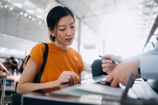 Woman in orange shirt writing on a form at an airport counter while another person hands her a pen