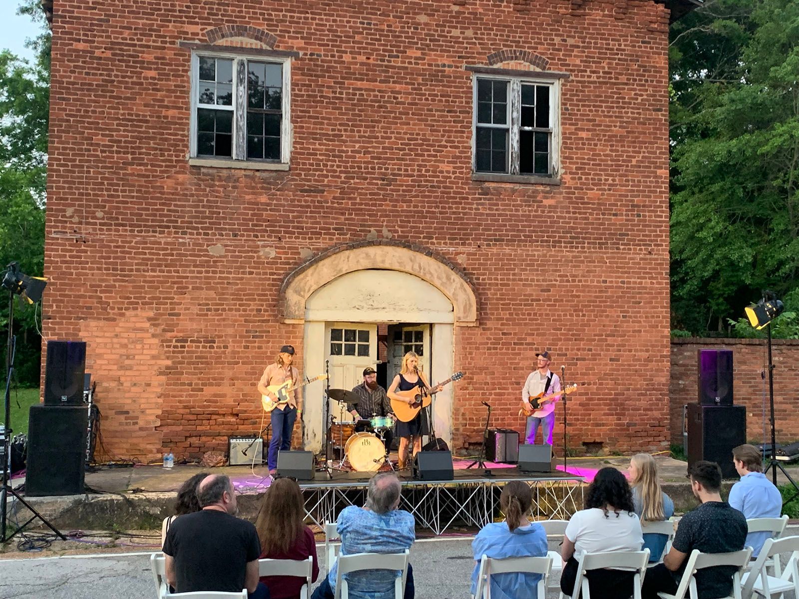 A group of people are sitting in chairs in front of a brick building watching a band perform on a stage