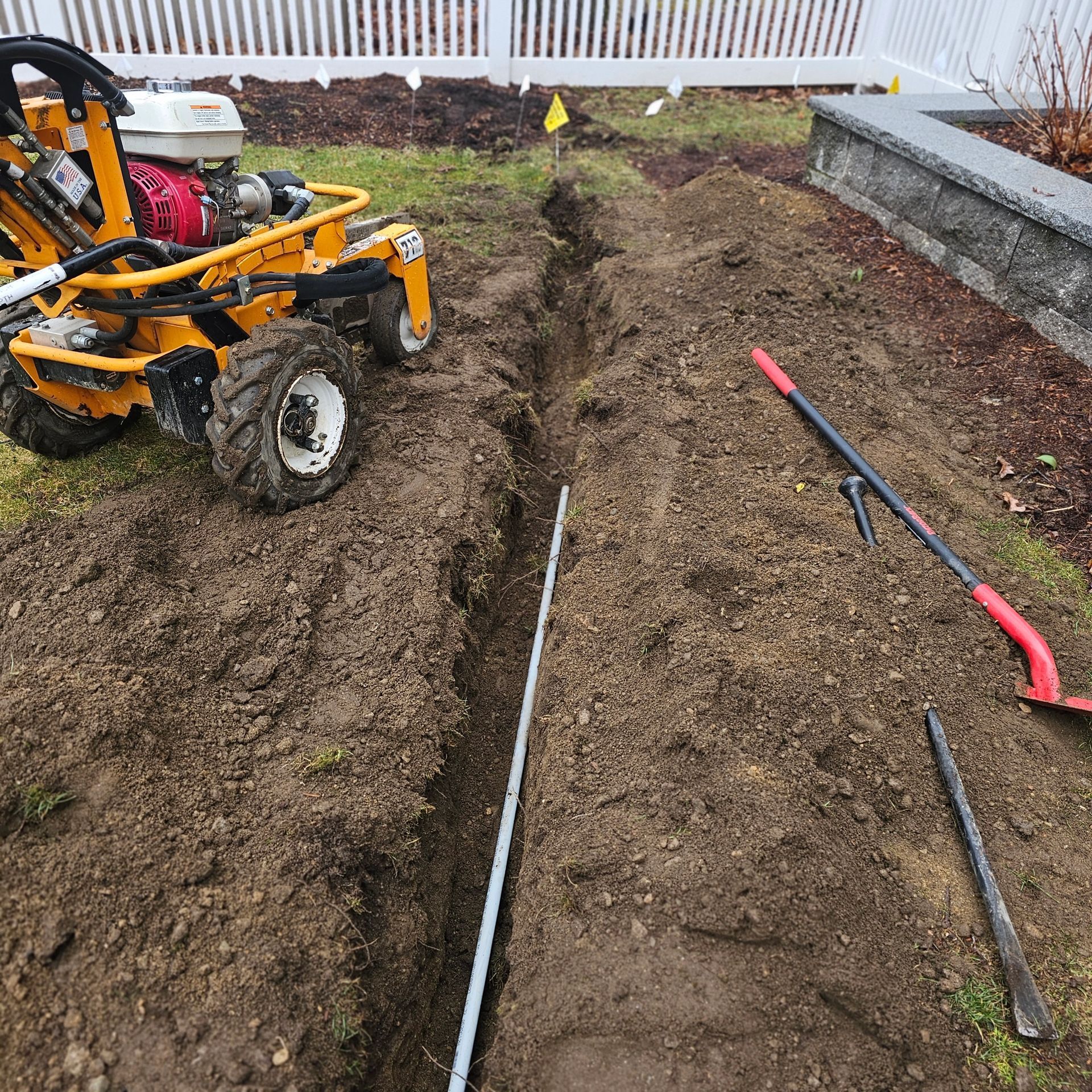 Trench dug in yard with trenching machine and tools. A white fence is in the background.
