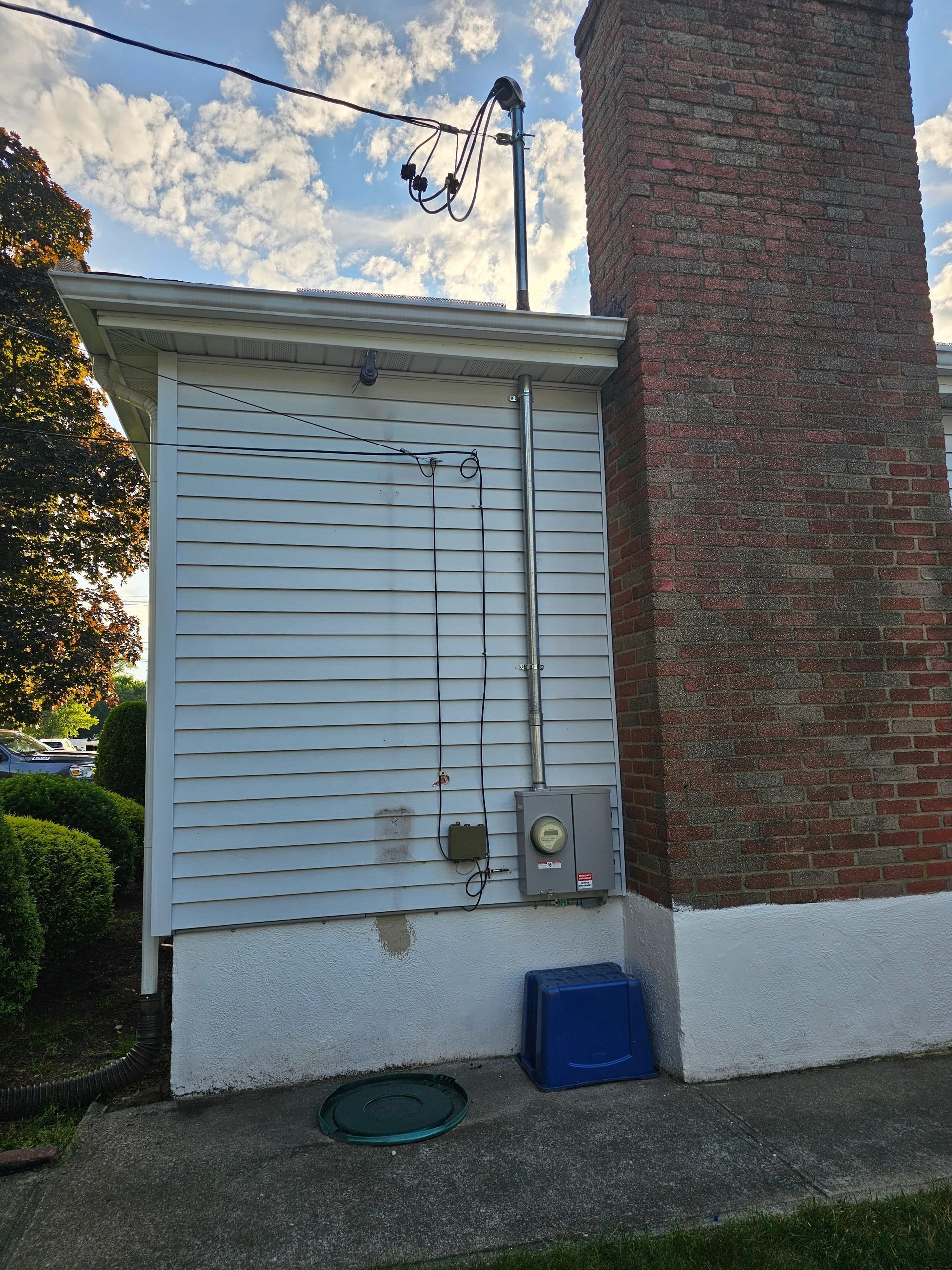 Side of a white building with electrical components and a brick chimney. A blue bin sits below.
