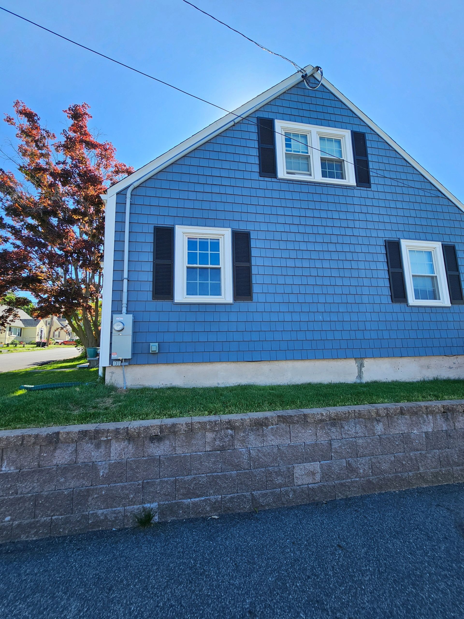 Blue house with white trim, black shutters, and a red tree under a sunny sky.