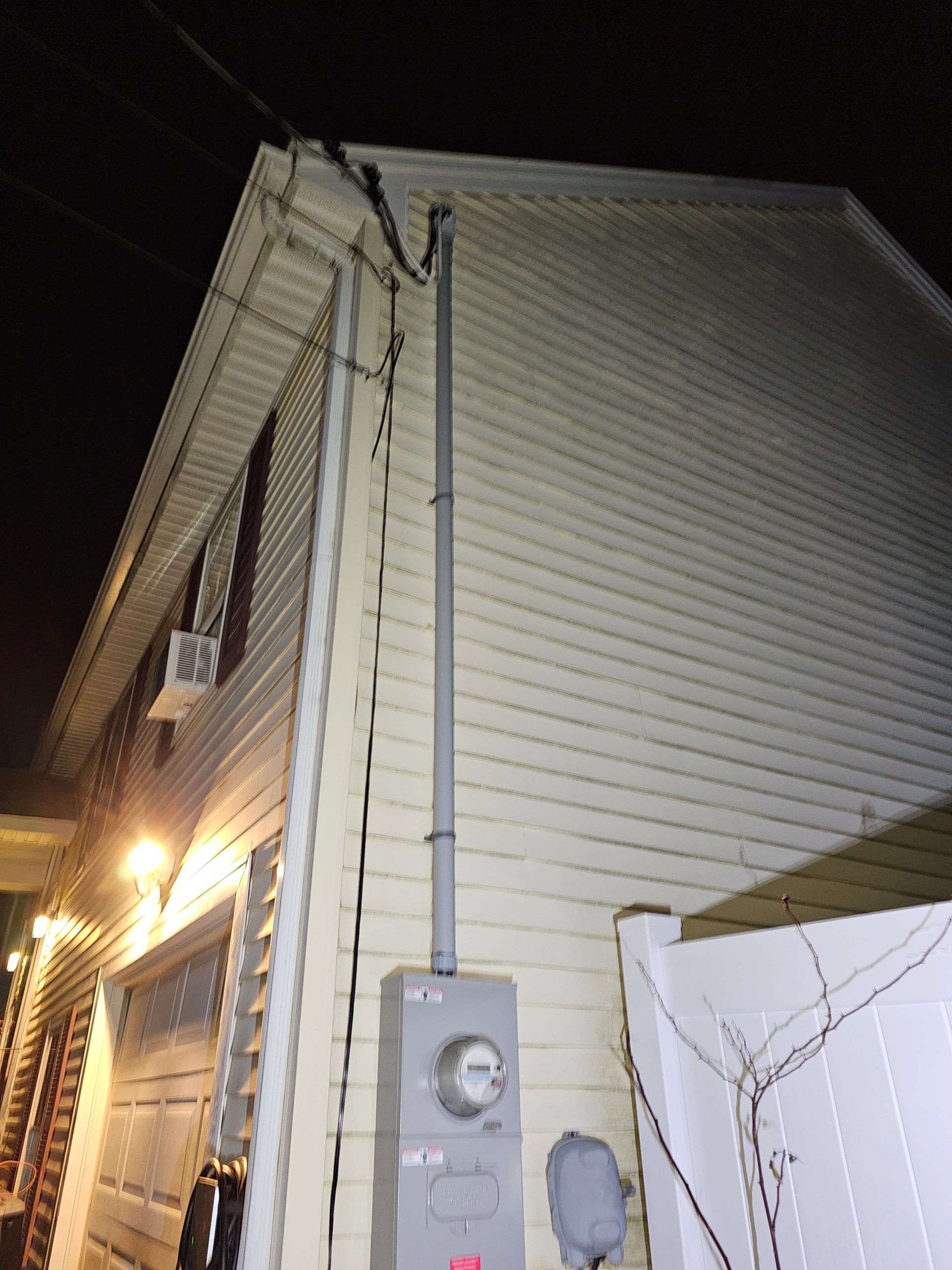 Exterior house with electrical meter, gray conduit, and utility wires against yellow siding at night.
