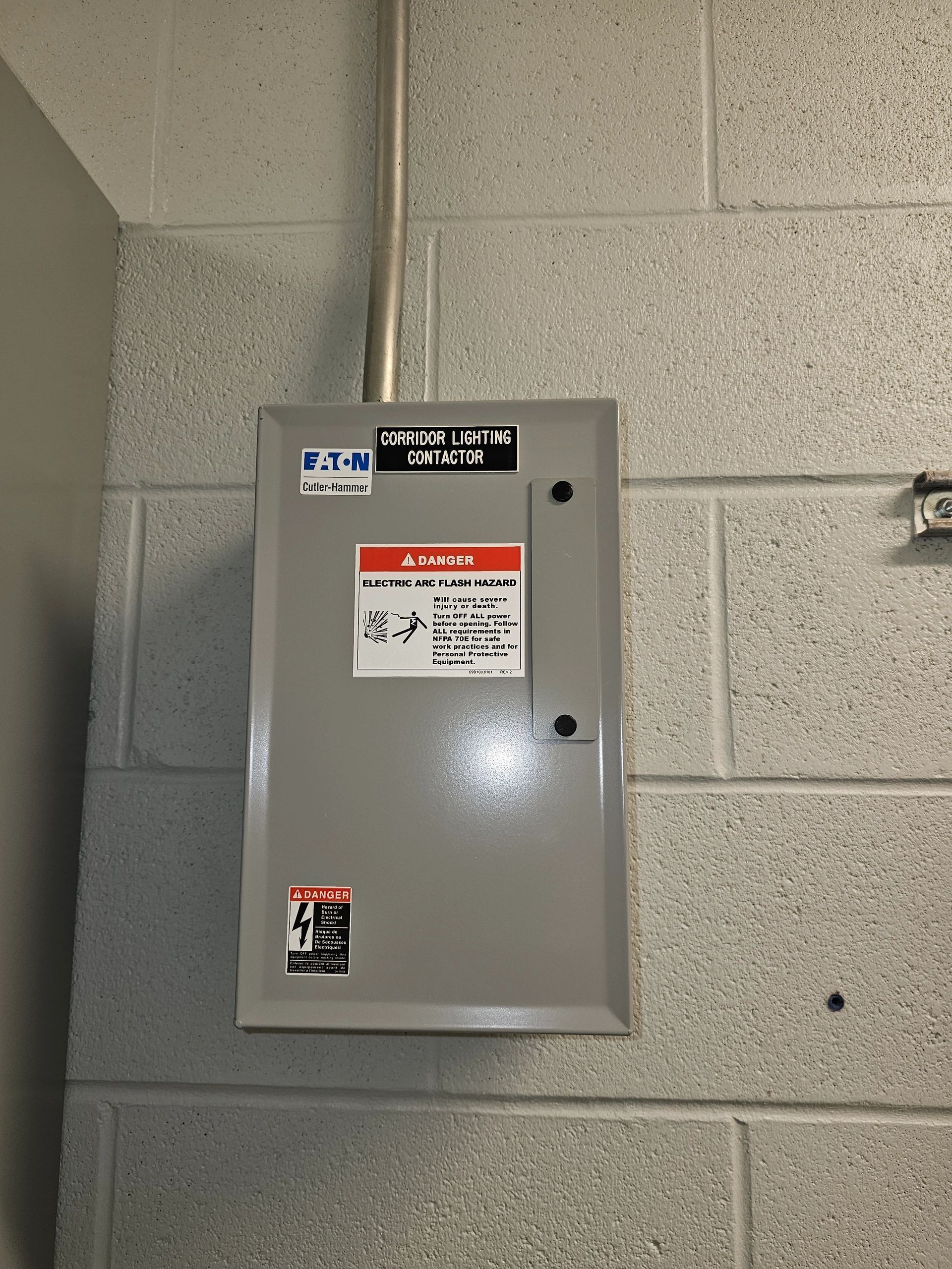 Gray electrical panel mounted on a cinder block wall, with warning labels and conduit running above.
