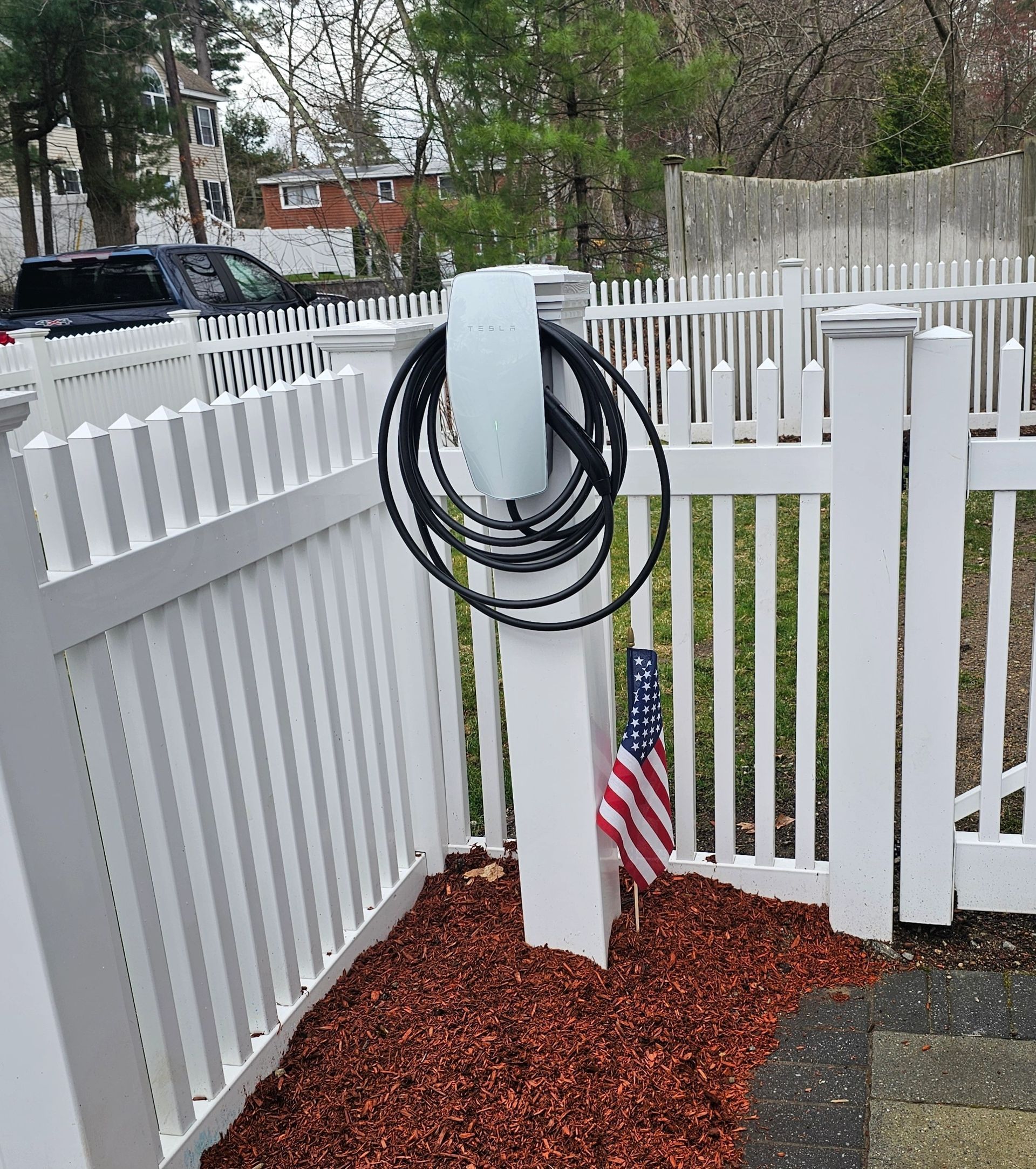Electric vehicle charger on a white picket fence post, cord coiled around it. American flag in front.