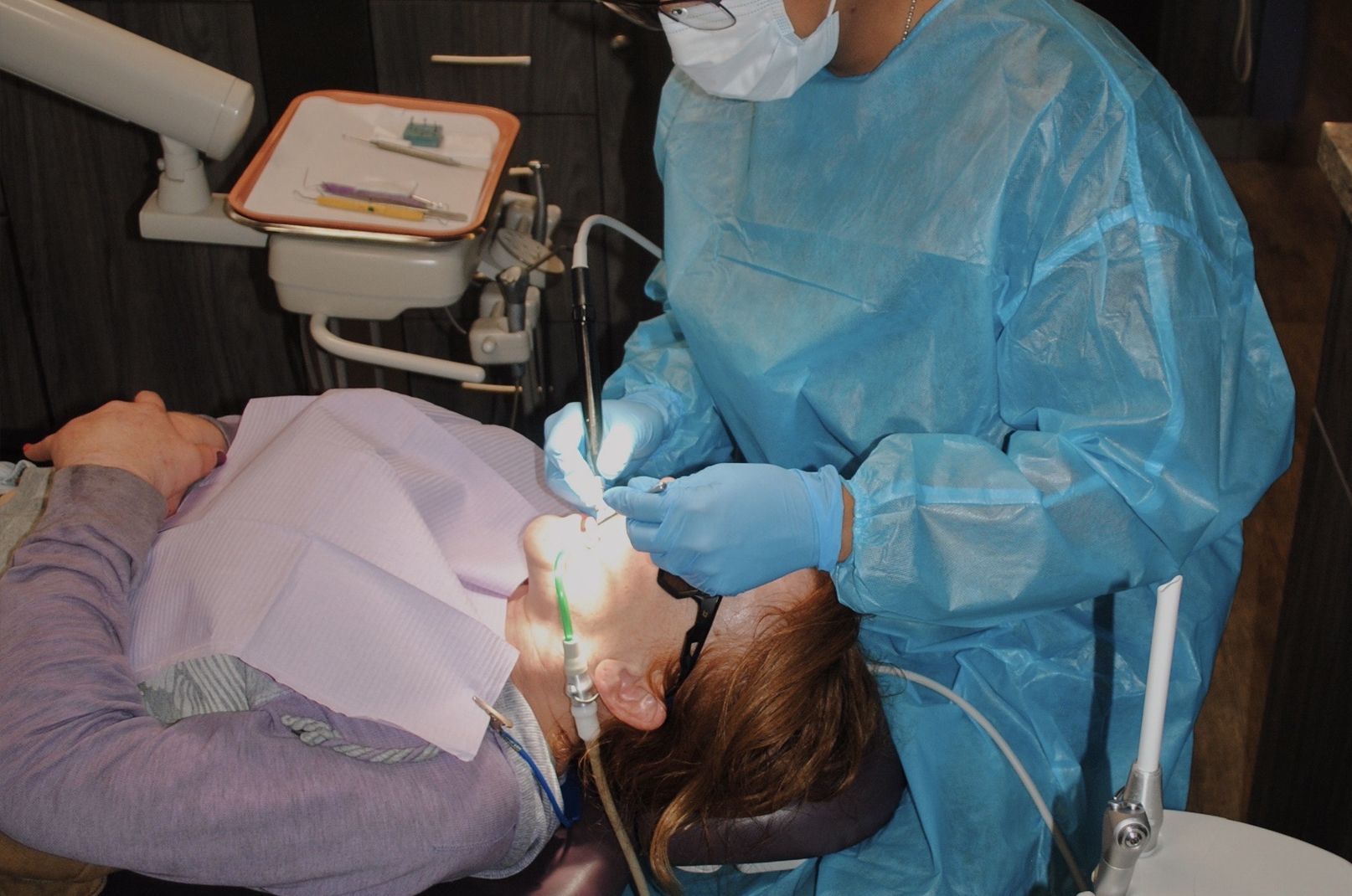 A dentist in blue scrubs is working on a patient in a dental chair.