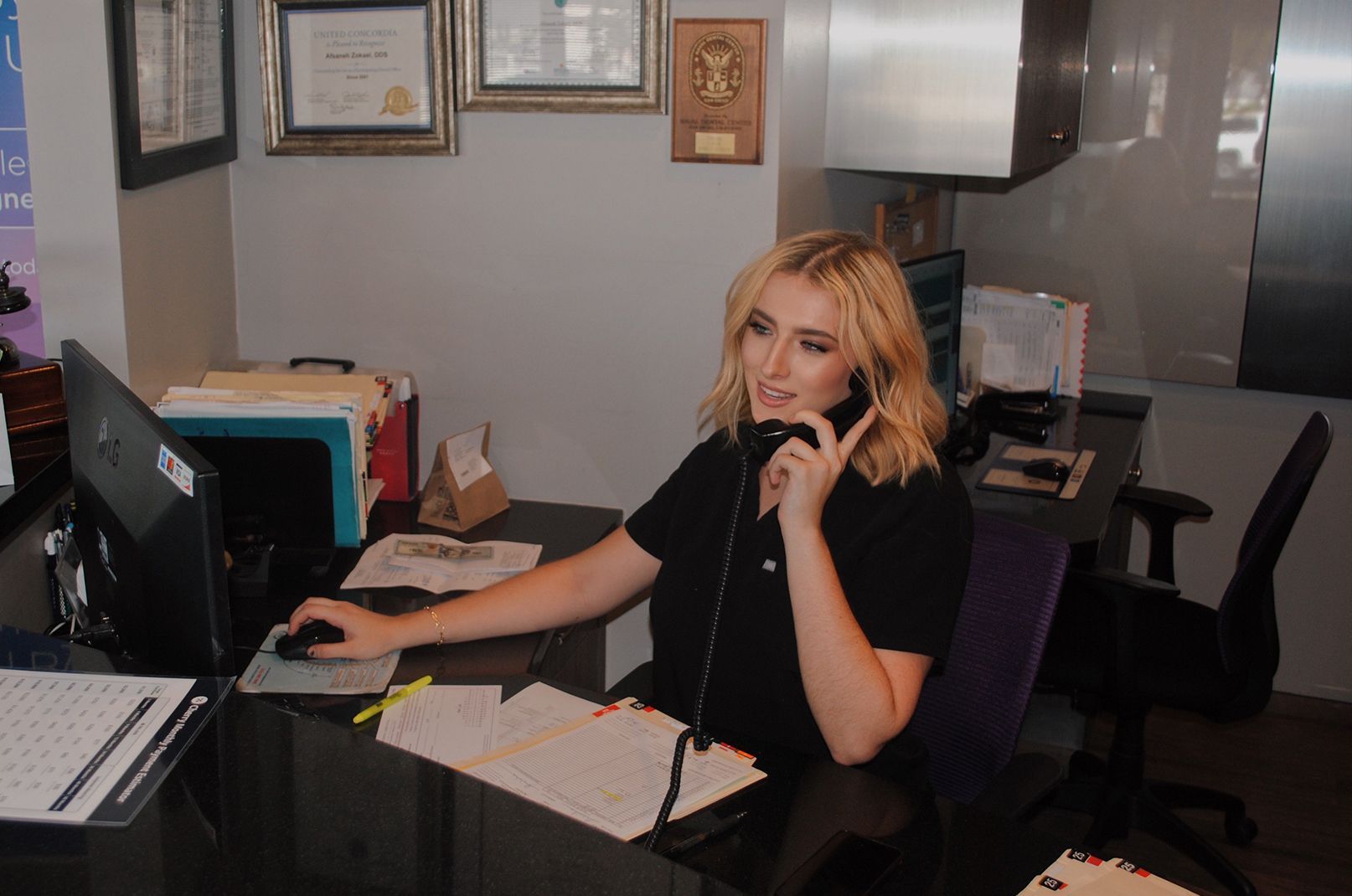 Woman at desk talking on phone, using computer, paperwork visible.