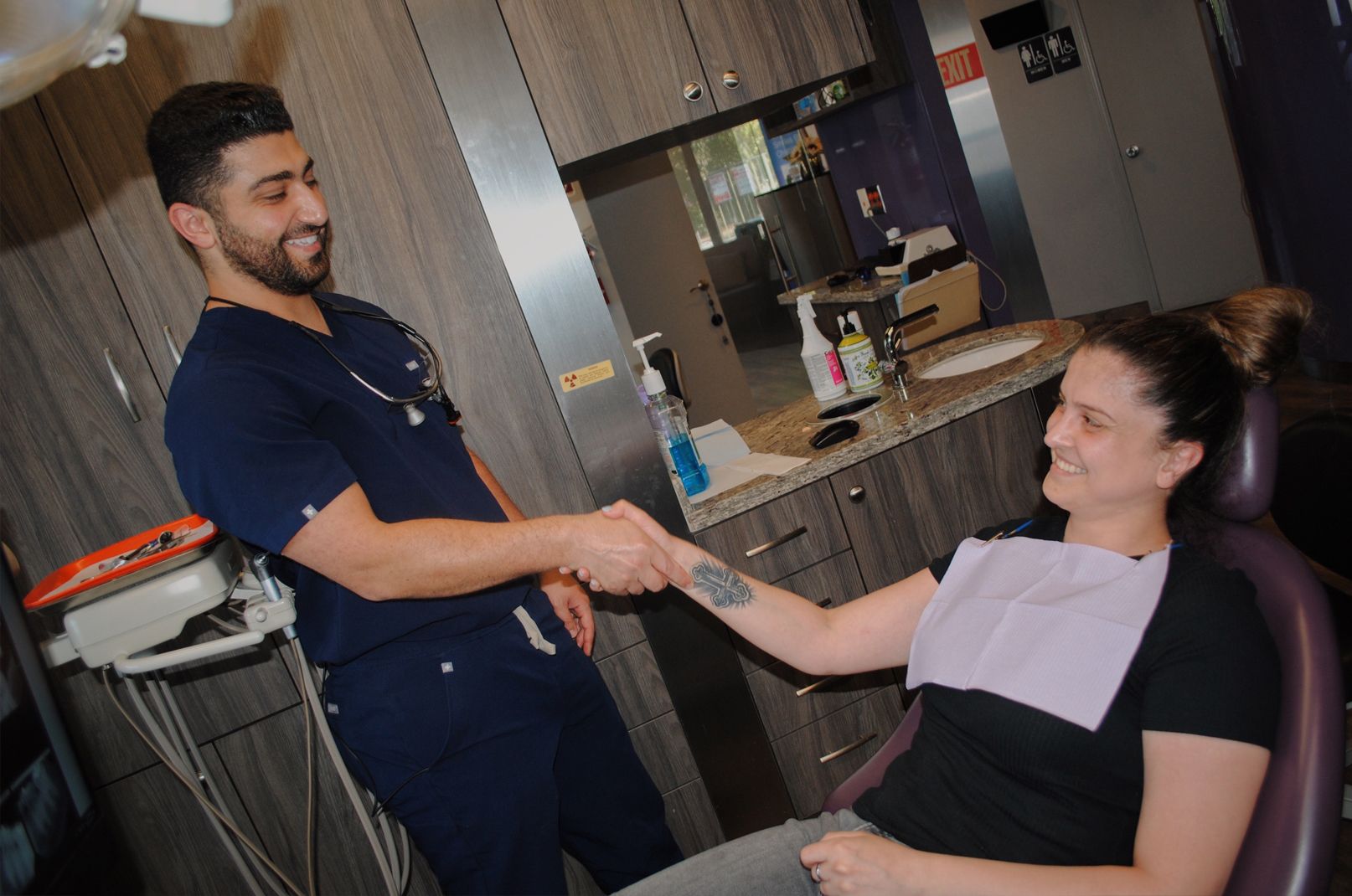 A dentist in blue scrubs shakes hands with a smiling patient in a dental chair.