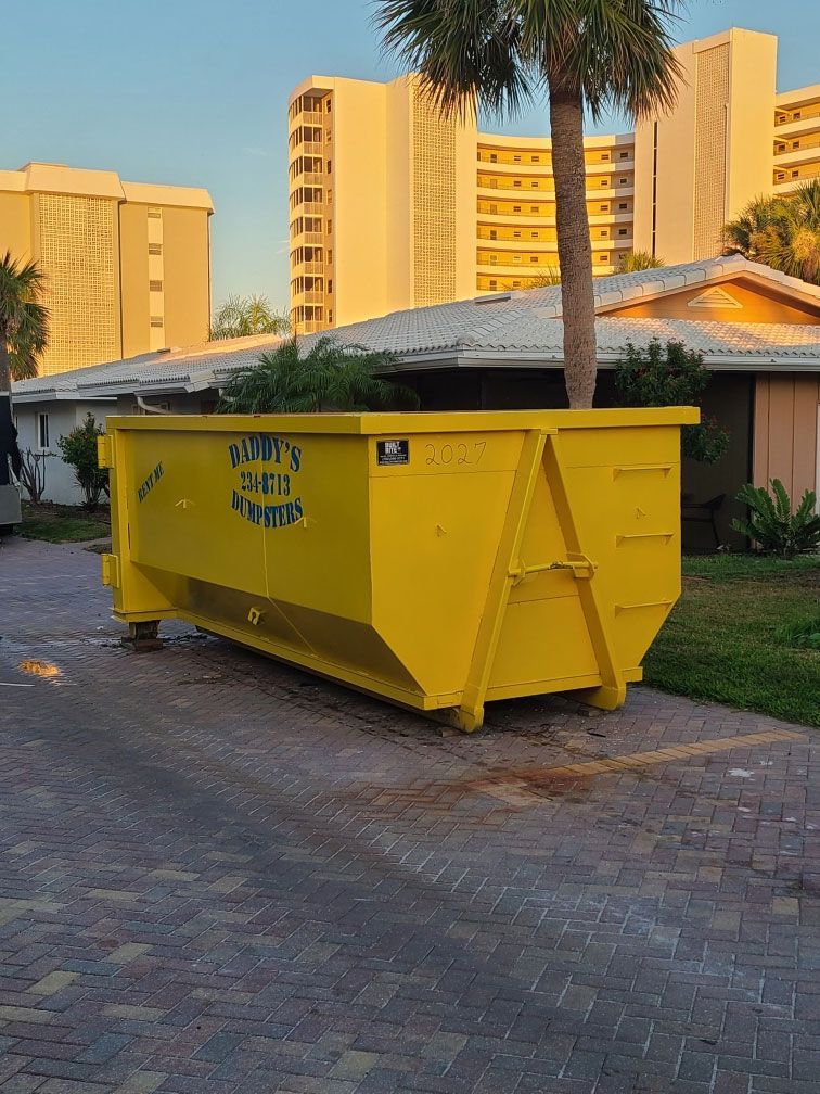 Yellow dumpster in front of a house with tall buildings in the background.