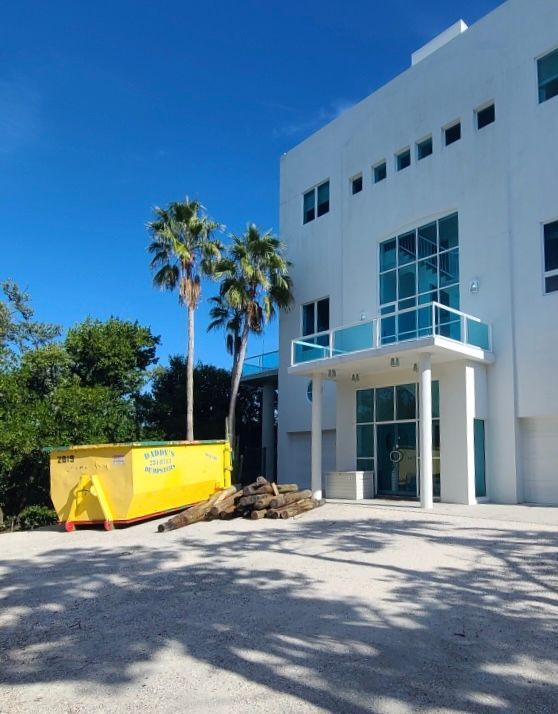 Yellow dumpster beside a white modern building, under a bright blue sky with palm trees.