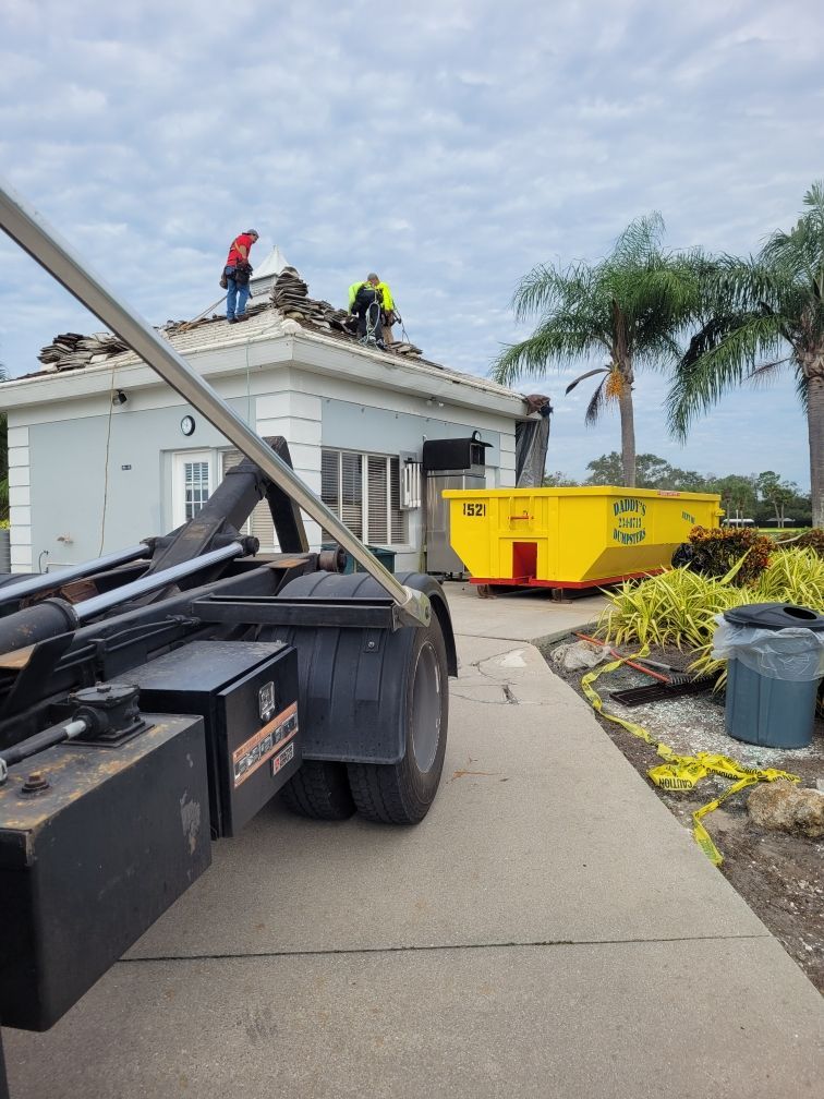 A dumpster is parked in front of a house with workers on the roof. Roll-Off Dumpster Rental in Sarasota.