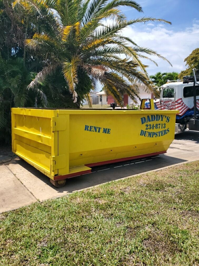 Yellow dumpster with Daddy's Dumpsters signage sits on pavement beside grass, with palm tree in background.