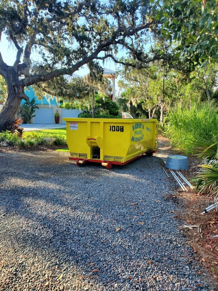 A yellow dumpster is parked in a gravel driveway next to a tree. Roll-Off Dumpster Rental in Sarasota.