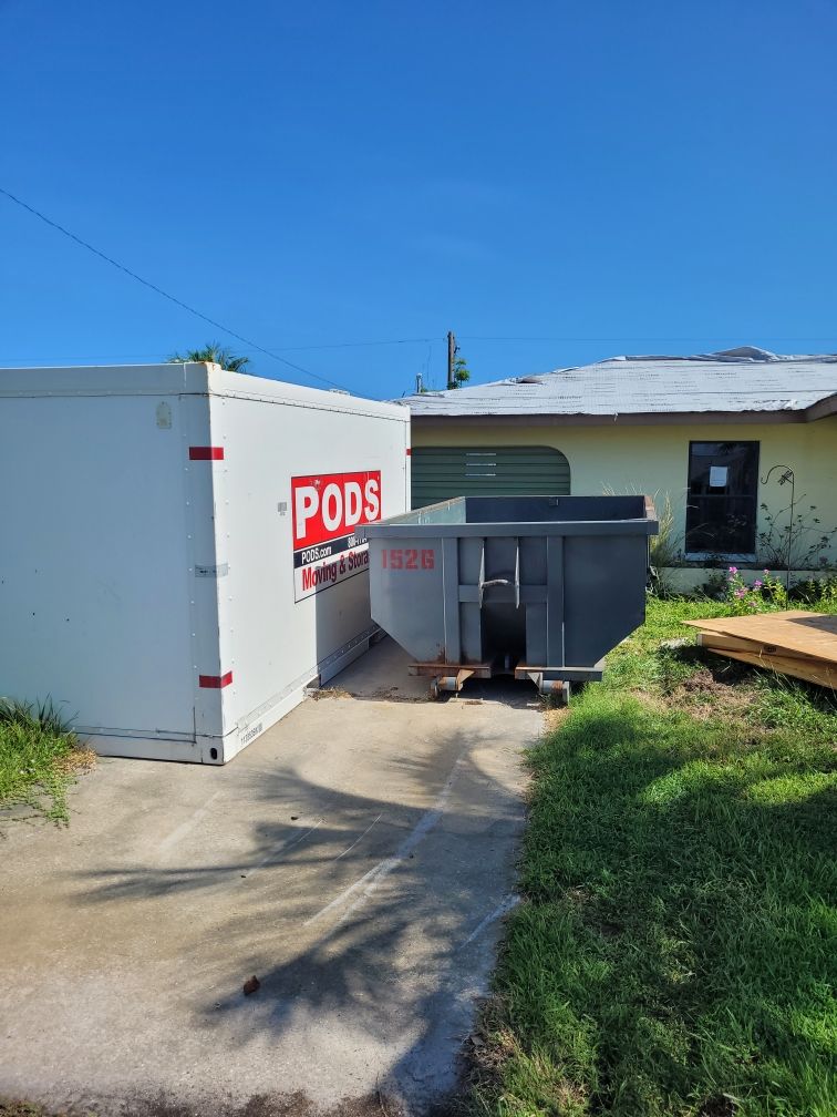 A yellow dumpster is parked in a gravel driveway next to a tree. Roll-Off Dumpster Rental in Sarasota.