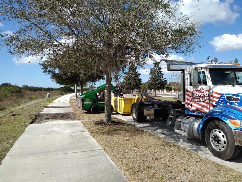 A row of trucks is parked on a sidewalk next to trees. Roll-Off Dumpster Rental in Sarasota.