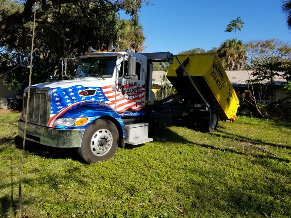 A dump truck with an American flag painted on it is parked in a grassy field. Roll-Off Dumpster Rental in Sarasota.
