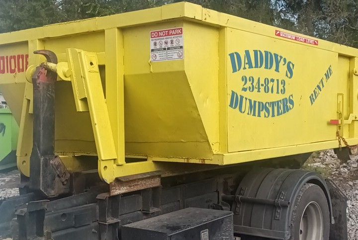A yellow dumpster truck is parked next to a green dumpster. Roll-Off Dumpster Rental in Sarasota.