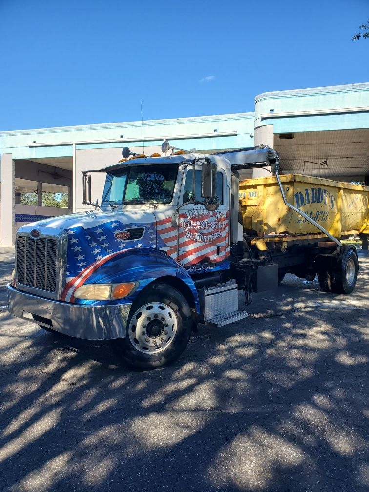 A dump truck with an American flag painted on the side is parked in front of a building. Roll-Off Dumpster Rental in Sarasota.