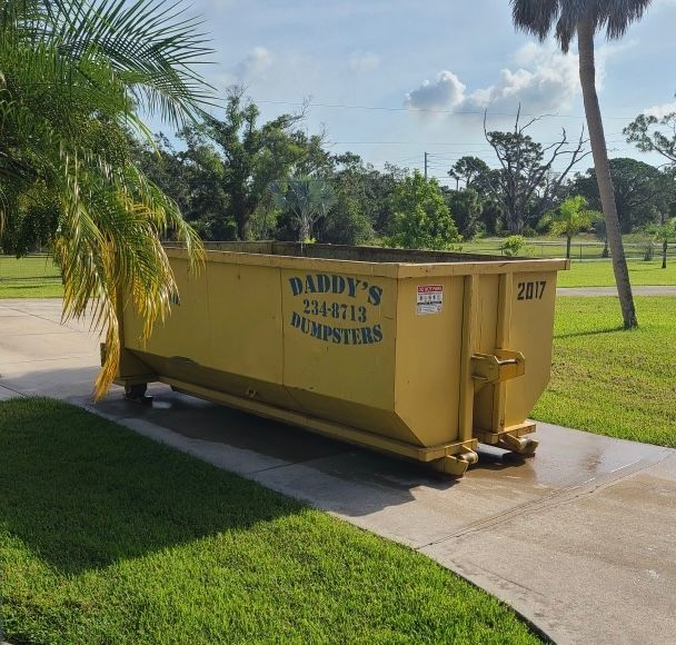 Yellow dumpster labeled 'Daddy's Dumpsters' on a driveway, near grass and palm trees.