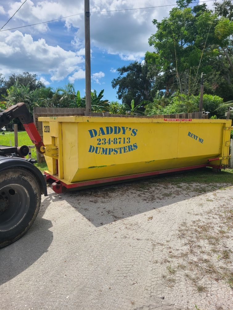 Yellow dumpster labeled 'Daddy's Dumpsters' on a driveway, next to a truck. Blue sky with clouds.