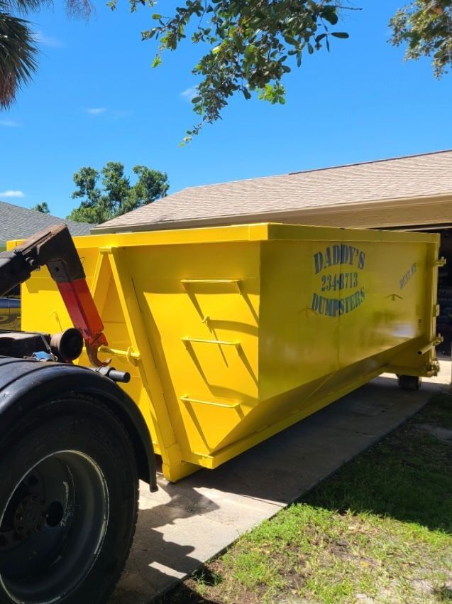 Yellow dumpster on a truck, parked in a driveway in front of a house on a sunny day.