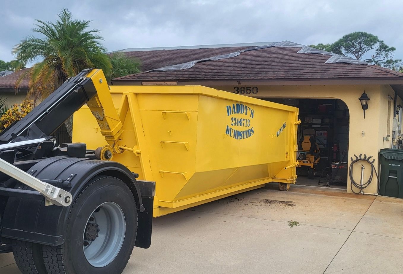 Yellow dumpster being placed in a garage by a truck. The garage is attached to a house with a brown roof.