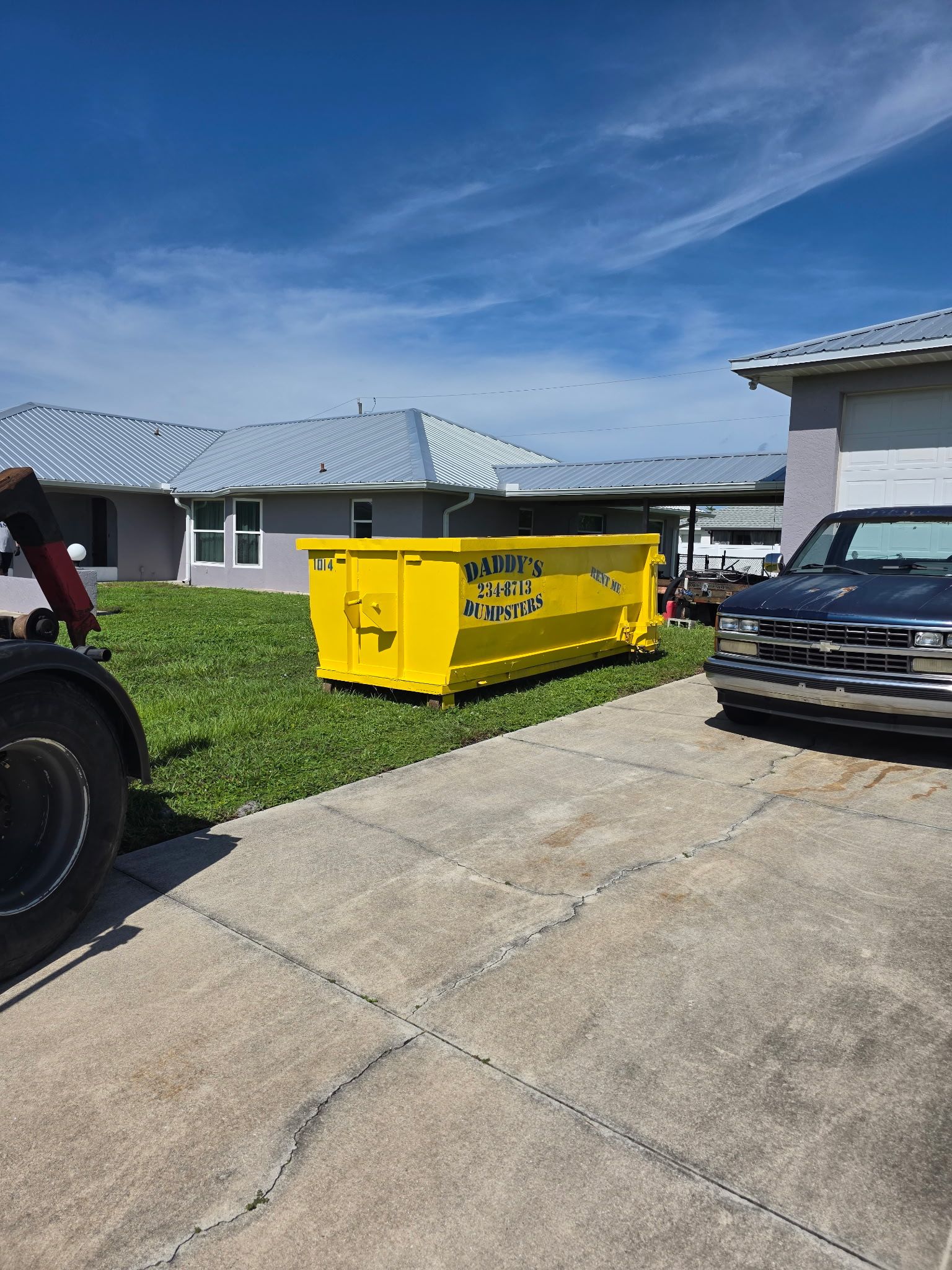 Bright yellow dumpster in a driveway beside parked vehicles under a blue sky