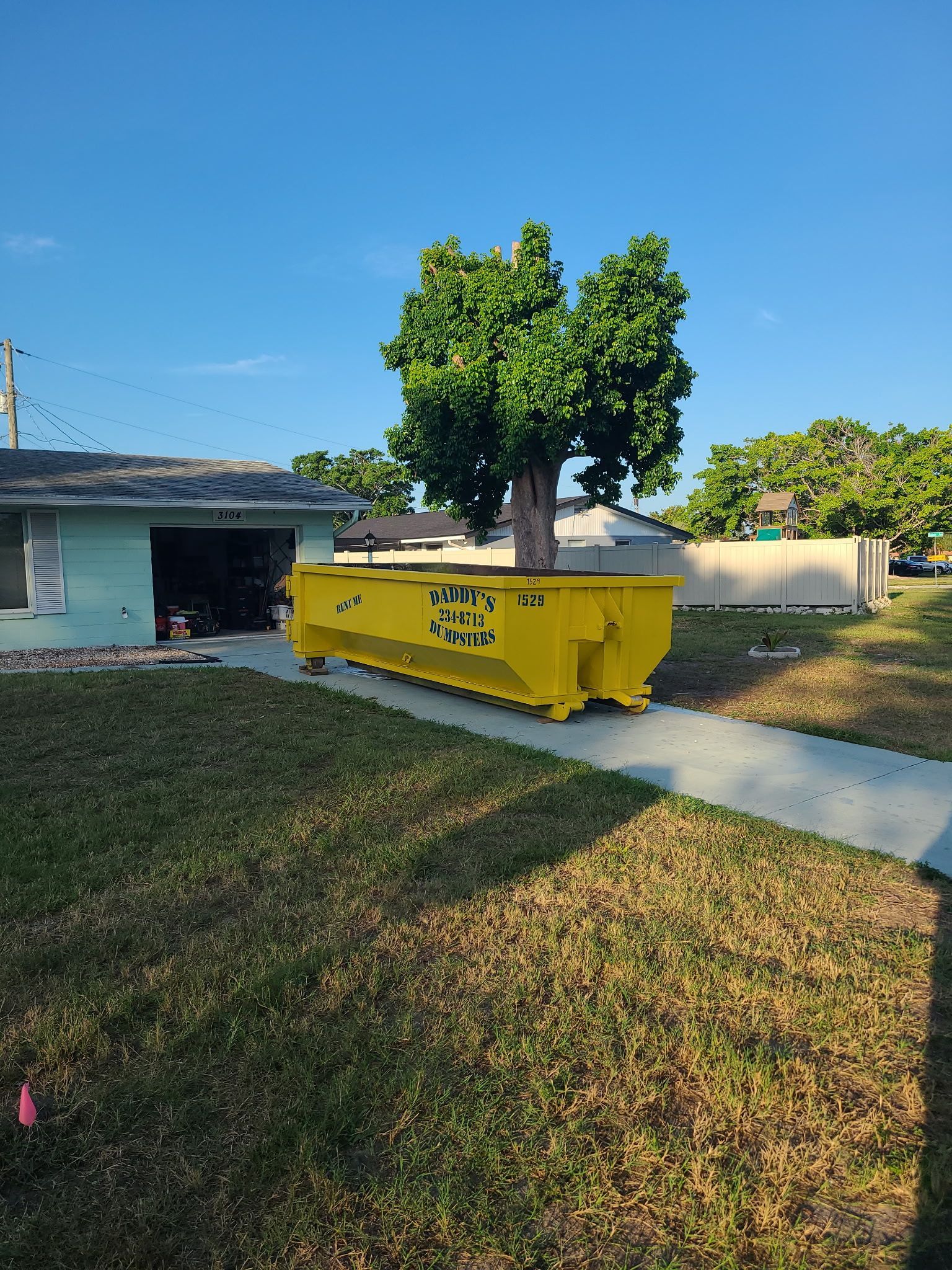 15-yard yellow dumpster beside a house on a sidewalk, with a tree and grassy yard under a clear blue sky