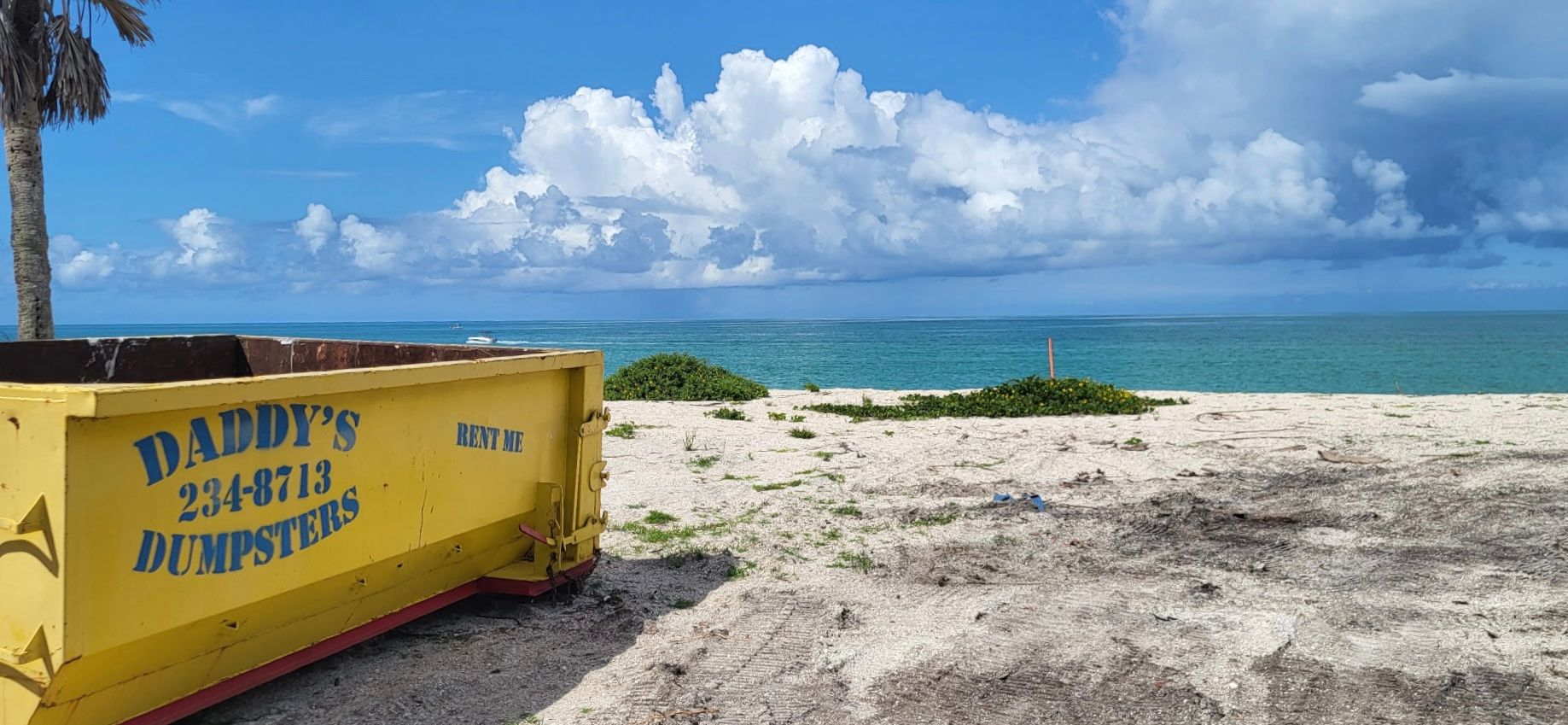 15-yard yellow dumpster on a sandy beach beside turquoise water under a bright cloudy sky