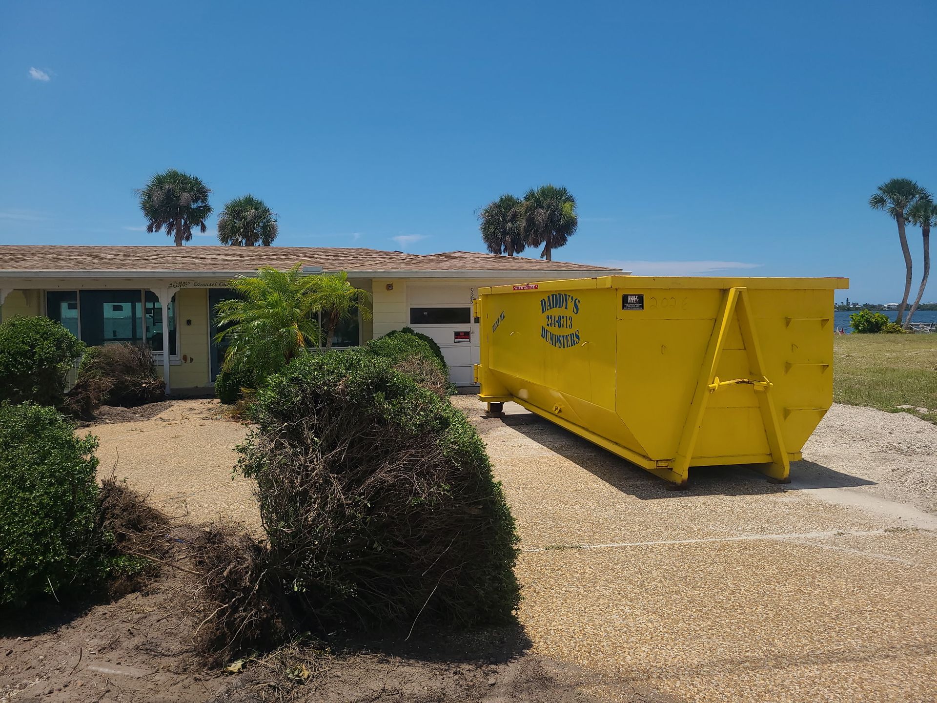 20-yard yellow dumpster in a sunny driveway beside a single-story house and shrubs.