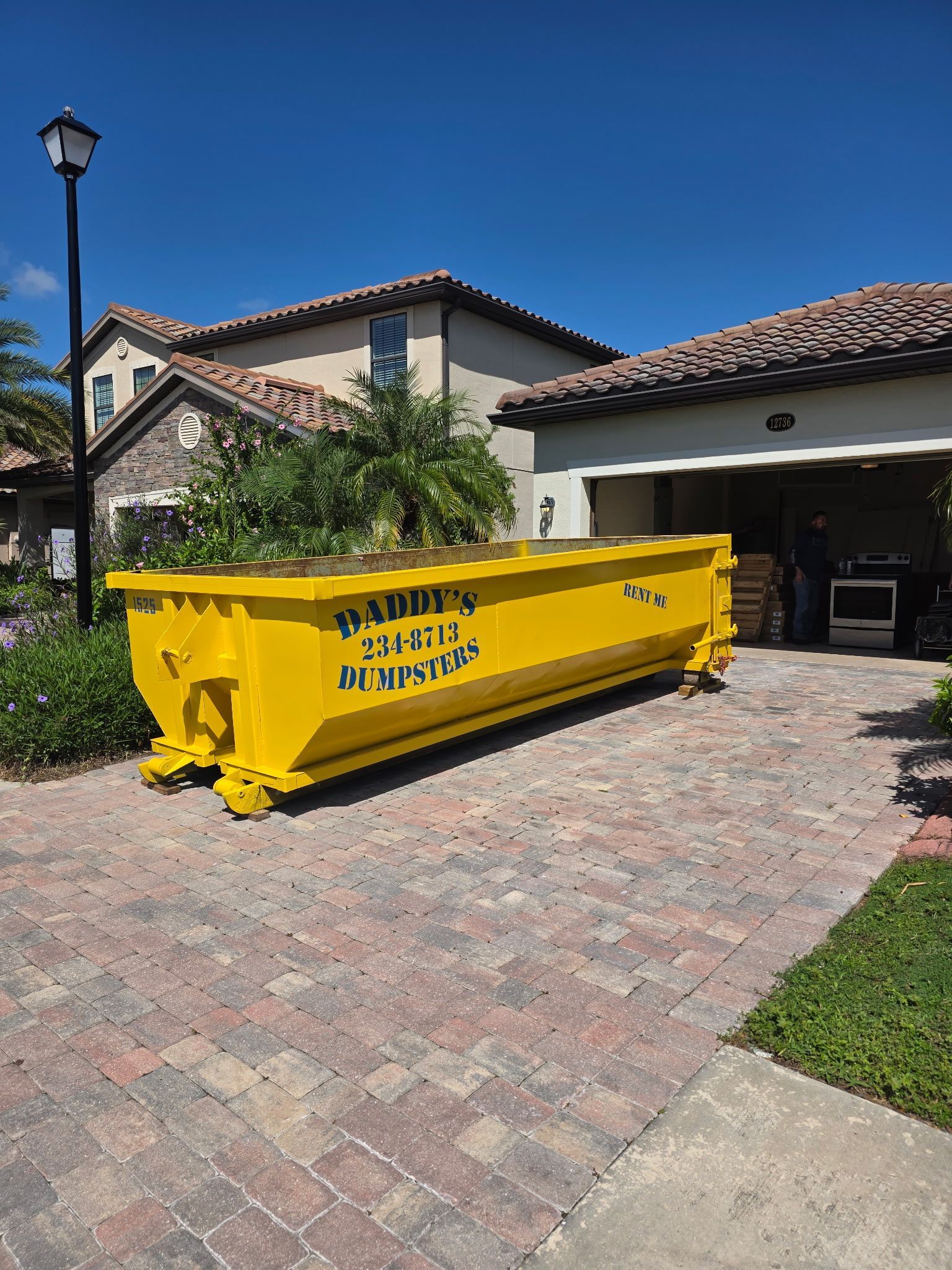 Yellow construction dumpster in a driveway in front of a house with a garage, on a brick paver surface.